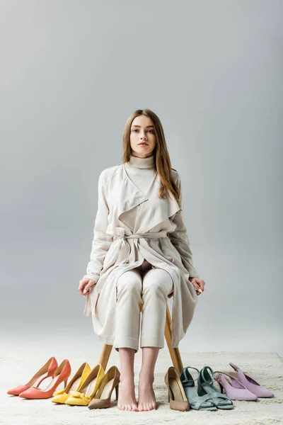 Stylish, confident girl looking at camera while sitting on chair near collection of shoes on grey background — Stock Photo