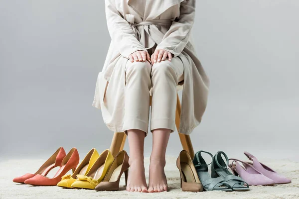 Vue recadrée de fille pieds nus assis sur la chaise près de la collection de chaussures sur fond gris — Photo de stock