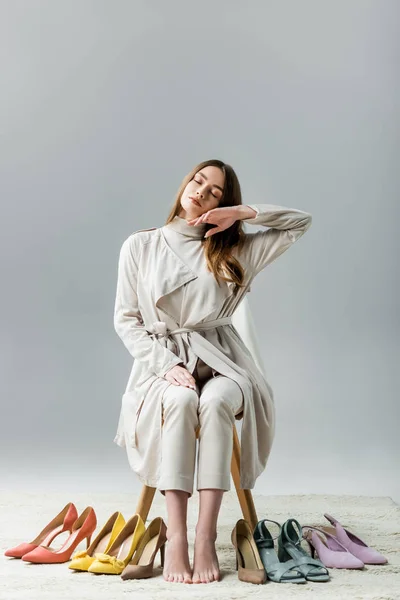 Chica cansada y elegante sentada en la silla cerca de la colección de zapatos y la cara conmovedora con la mano sobre fondo gris - foto de stock