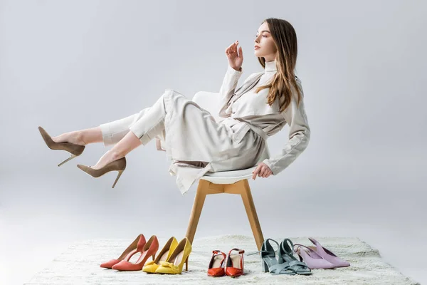 Thoughtful girl sitting on chair with raised legs near collection of shoes and looking away on grey background — Stock Photo