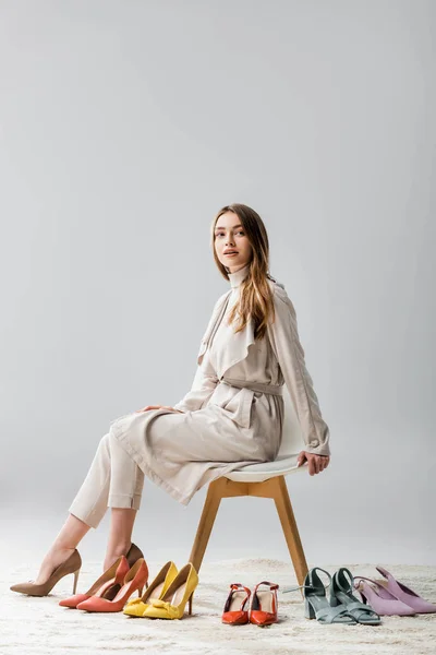 Elegante mujer joven mirando hacia otro lado mientras está sentado en la silla cerca de la colección de zapatos sobre fondo gris - foto de stock