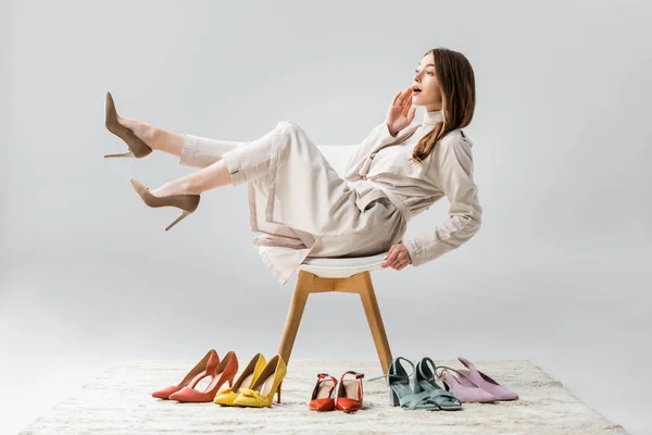 Surprised girl sitting on chair with raised legs and looking away near collection of shoes on grey background — Stock Photo