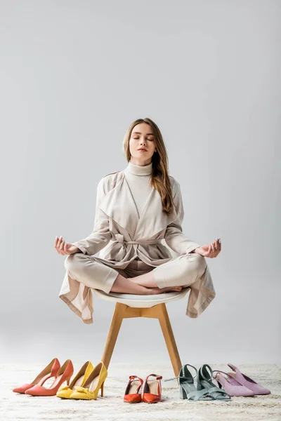 Élégante fille assise sur une chaise en lotus pose près de la collection de chaussures sur fond gris — Photo de stock