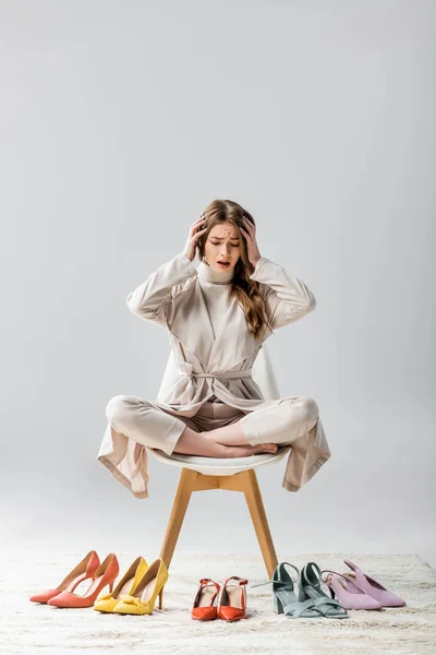 Chica impactada sentada en silla en pose de loto, mirando la colección de zapatos y tocando la cabeza sobre fondo gris - foto de stock