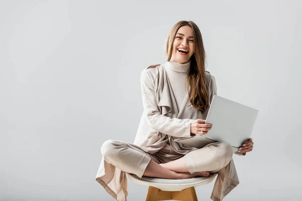 Fröhliches Mädchen blickt in die Kamera, während es auf einem Stuhl in Lotus-Pose sitzt und Laptop isoliert auf grau hält — Stockfoto