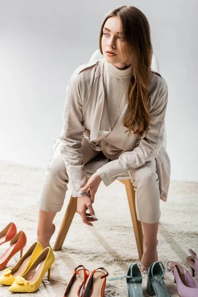 Ragazza elegante guardando lontano mentre seduto vicino collezione di scarpe su sfondo grigio — Foto stock