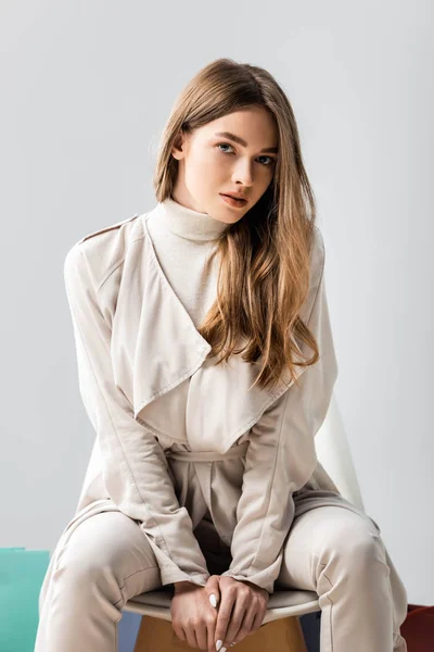 Menina atraente e elegante olhando para a câmera enquanto sentada na cadeira isolada no cinza — Fotografia de Stock