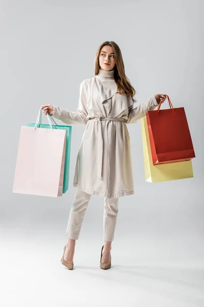 Vista completa de la chica con estilo mirando hacia otro lado mientras sostiene bolsas de compras sobre fondo gris - foto de stock
