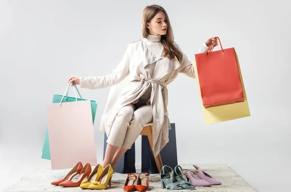 Chica de moda sentado en la silla cerca de zapatos en el suelo y la celebración de bolsas de compras en fondo gris - foto de stock