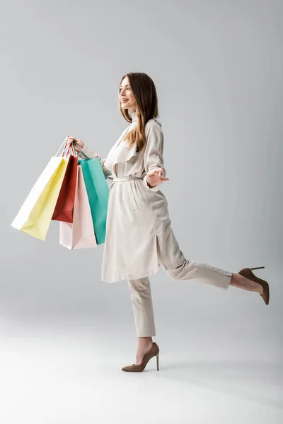 Visão de comprimento total da menina feliz na capa elegante segurando sacos de compras no fundo cinza — Fotografia de Stock