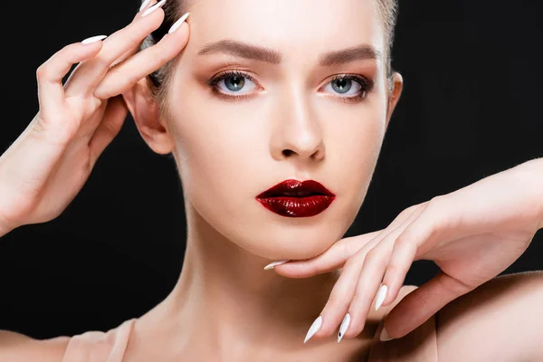Attractive young woman with red lips and makeup touching face while looking at camera isolated on black — Stock Photo