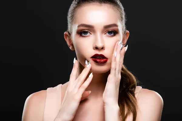 Bonita mujer joven con labios rojos y maquillaje tocando la cara mientras mira la cámara aislada en negro - foto de stock