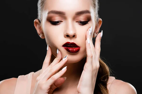 Atractiva joven con labios rojos y maquillaje tocando la cara aislado en negro - foto de stock
