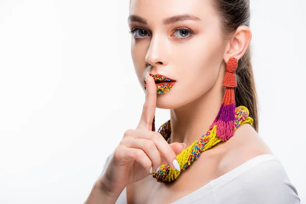 Attractive girl in beaded accessories, with beads on lips, showing hush sign and looking at camera isolated on white — Stock Photo