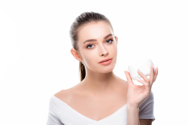 Chica atractiva mirando a la cámara mientras sostiene contenedor con crema facial aislado en blanco - foto de stock