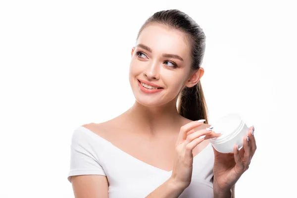 Привлекательная, веселая девушка смотрит в сторону, касаясь крема для лица в контейнере, изолированном на белом — стоковое фото