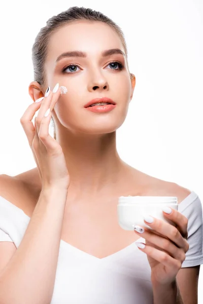 Hermosa, mujer sonriente aplicando crema cosmética en la cara y mirando hacia otro lado aislado en blanco - foto de stock