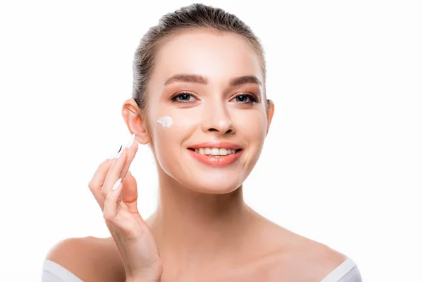 Atractiva, mujer sonriente aplicando crema cosmética en la cara y mirando a la cámara aislada en blanco - foto de stock