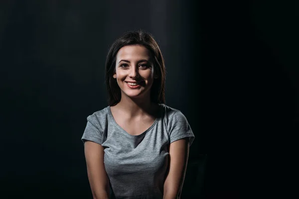 Happy young woman smiling at camera while sitting in darkness isolated on black — Stock Photo