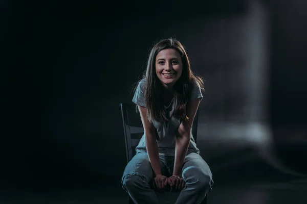 Happy young woman smiling at camera while sitting in darkness on black background — Stock Photo