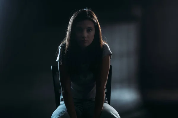 Hopeless young woman looking at camera while sitting on chair in darkness on black background — Stock Photo