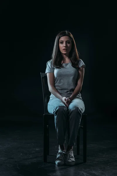 Sad young woman looking at camera while sitting on chair in darkness on black background — Stock Photo