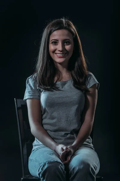 Cheerful young woman looking at camera while sitting in darkness isolated on black — Stock Photo