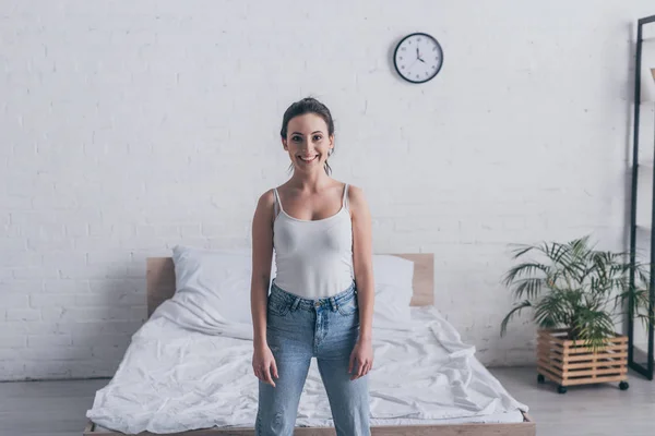 Feliz joven mujer de pie en el dormitorio y sonriendo a la cámara - foto de stock