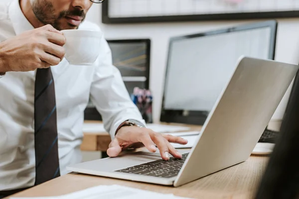 Cropped view of bi-racial trader using laptop and drinking coffee — Stock Photo