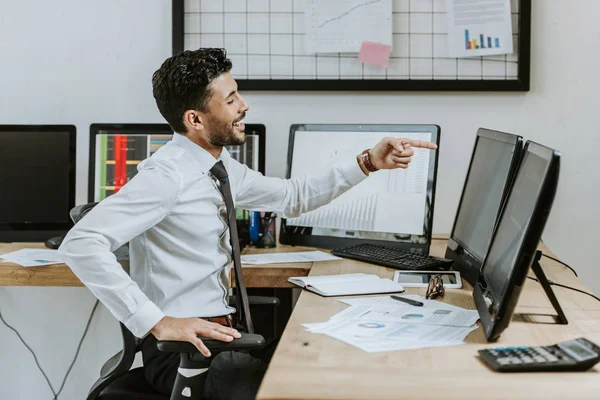 Vista lateral del comerciante bi-racial sonriente apuntando con el dedo a la computadora - foto de stock