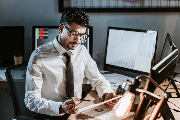 Handsome bi-racial trader in glasses looking at paper and sitting at table — Stock Photo