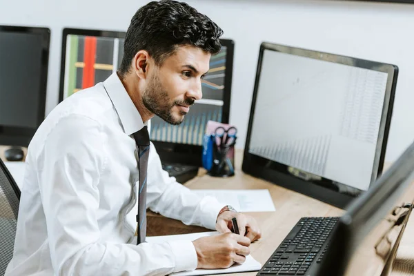 Commerçant bi-racial regardant l'ordinateur et tenant stylo dans le bureau — Photo de stock