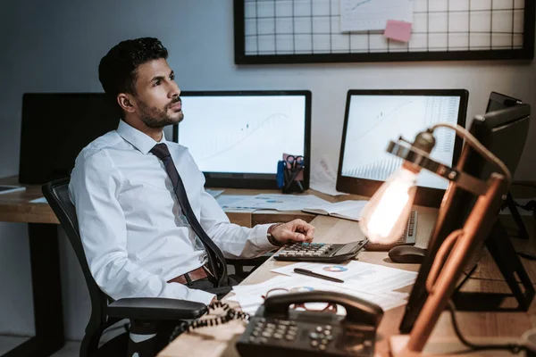 Pensoso commerciante bi-razziale seduto vicino ai computer e guardando altrove — Foto stock