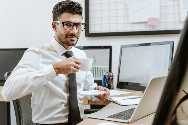 Sonriente bi-racial comerciante sosteniendo taza con café y sentado cerca de computadoras - foto de stock