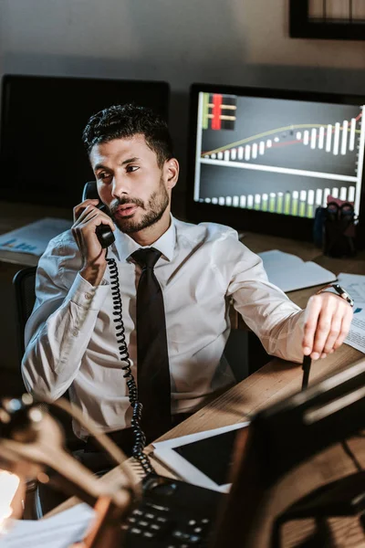 Vue grand angle du commerçant bi-racial parlant au téléphone et assis près de l'ordinateur avec des graphiques — Photo de stock