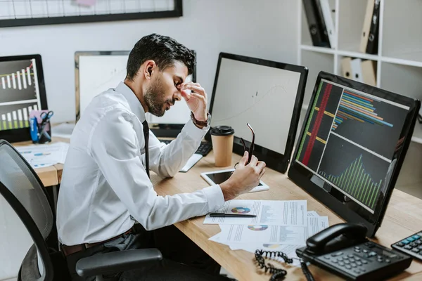 Vista lateral do comerciante bi-racial cansado sentado perto de computadores com gráficos e segurando óculos — Fotografia de Stock