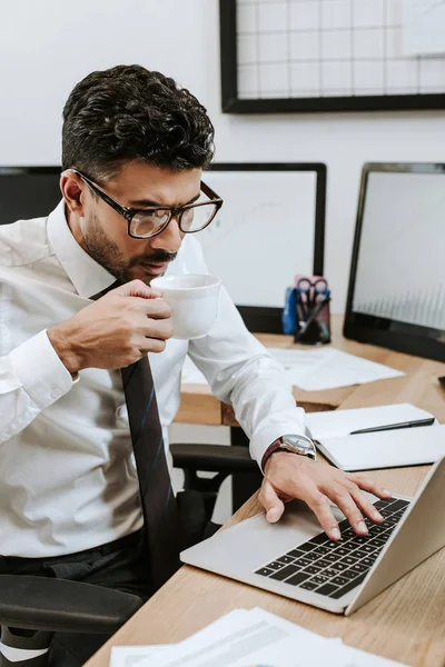Bello bi-razziale trader in occhiali utilizzando il computer portatile e bere caffè — Foto stock