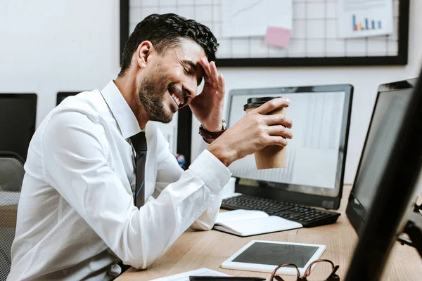 Sorrindo comerciante bi-racial segurando copo de papel e sentado perto do computador — Fotografia de Stock