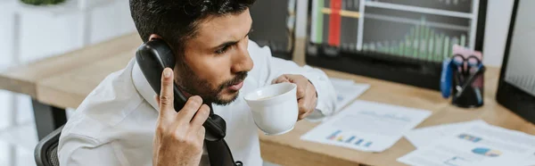 Panoramic shot of bi-racial trader drinking coffee and talking on telephone — Stock Photo