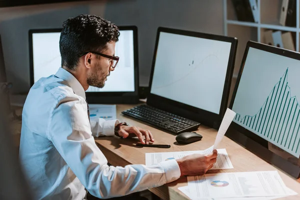 Commerçant bi-racial regardant l'ordinateur avec des graphiques et tenant du papier — Stock Photo