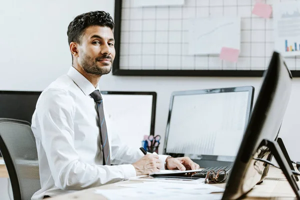 Lächelnder Händler zwischen den Rassen, der am Tisch sitzt und neben Computern sitzt — Stockfoto