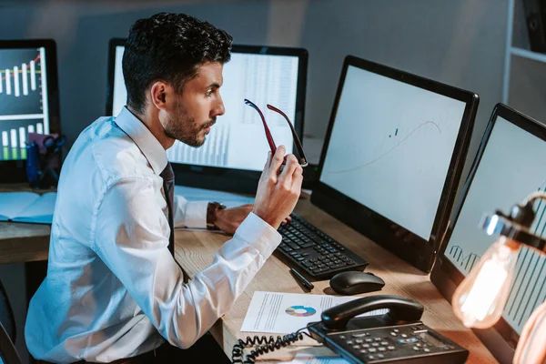 Visão lateral do comerciante bi-racial segurando óculos e olhando para o computador com gráficos — Fotografia de Stock