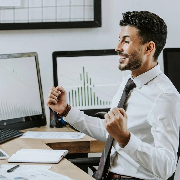 Seitenansicht eines lächelnden bi-rassischen Händlers, der Ja-Geste zeigt und neben Computern sitzt — Stockfoto