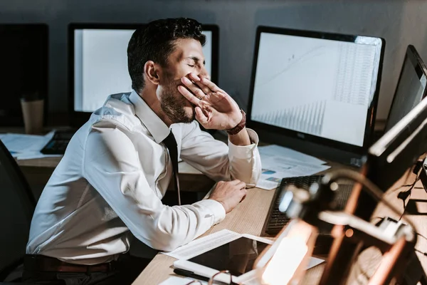 Comerciante bi-racial bostezando y sentado cerca de la computadora con gráficos - foto de stock