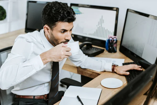 Vista de ángulo alto de bi-racial comerciante beber café y mirando a la computadora - foto de stock