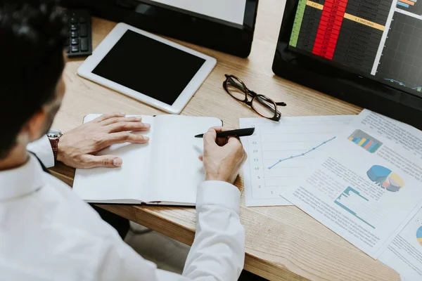 Vista ritagliata di bi-razziale trader scrittura in notebook e seduto a tavola — Foto stock