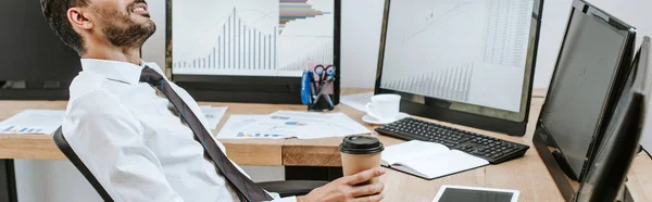 Panoramaaufnahme eines lächelnden Händlers, der Pappbecher in der Hand hält und neben Computern mit Grafiken sitzt — Stockfoto