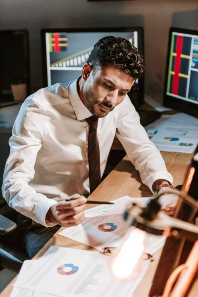 Zwei-Rassen-Händler, der Papier betrachtet und neben Computern mit Grafiken sitzt — Stockfoto