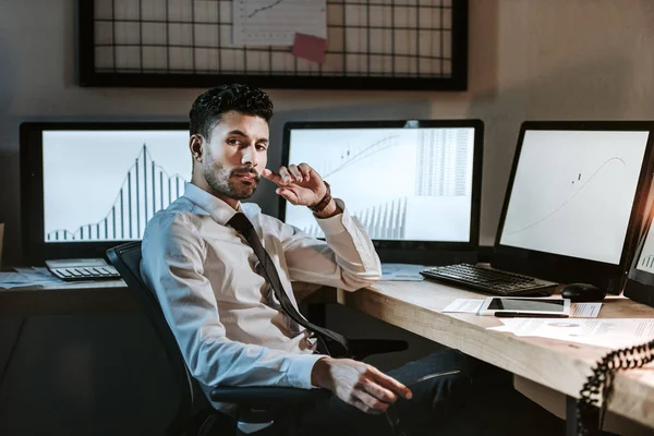 Nachdenklicher bi-rassischer Händler, der neben Computern mit Grafiken sitzt und in die Kamera blickt — Stock Photo