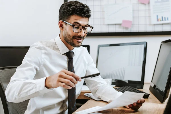 Comerciante bi-racial sonriente sentado en la mesa y mirando el papel - foto de stock